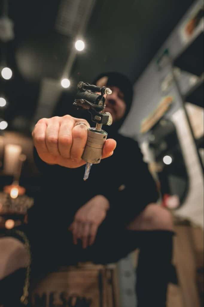 Tattoo artist_ Tattoo artist holding a tattoo machine in focus, with a blurred studio backdrop. | Sky Rye Design Tattoo artist holding a tattoo machine in focus, with a blurred studio backdrop.