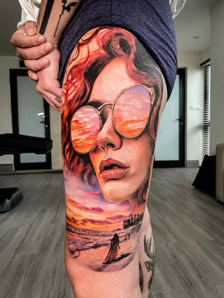 Tattoo design on thigh depicting woman in sunglasses, reflection shows a vibrant sunset beach scene.