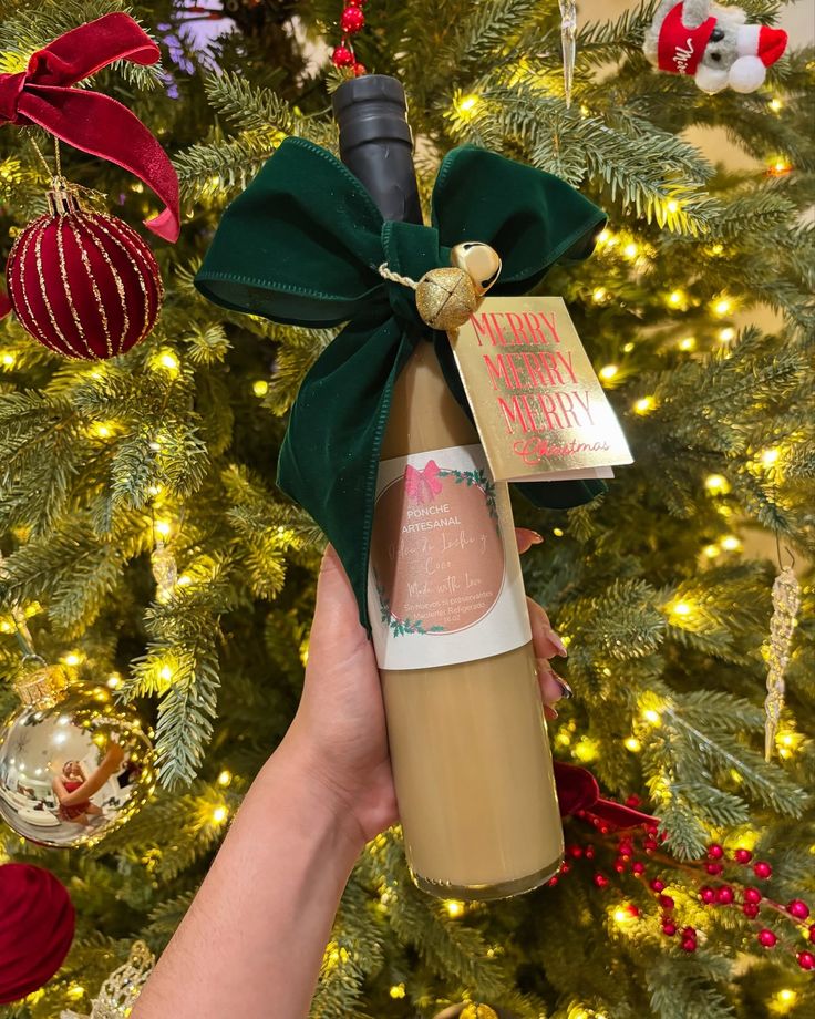 Te gustaría disfrutar del cremoso y delicioso sabor del dulce de leche y coco con toques de ron dorado _ __Ideal para compartir con familiares y amigos ❤️ Festive eggnog bottle with green bow and holiday tag, held in front of a beautifully decorated Christmas tree. | Sky Rye Design Festive eggnog bottle with green bow and holiday tag, held in front of a beautifully decorated Christmas tree.
