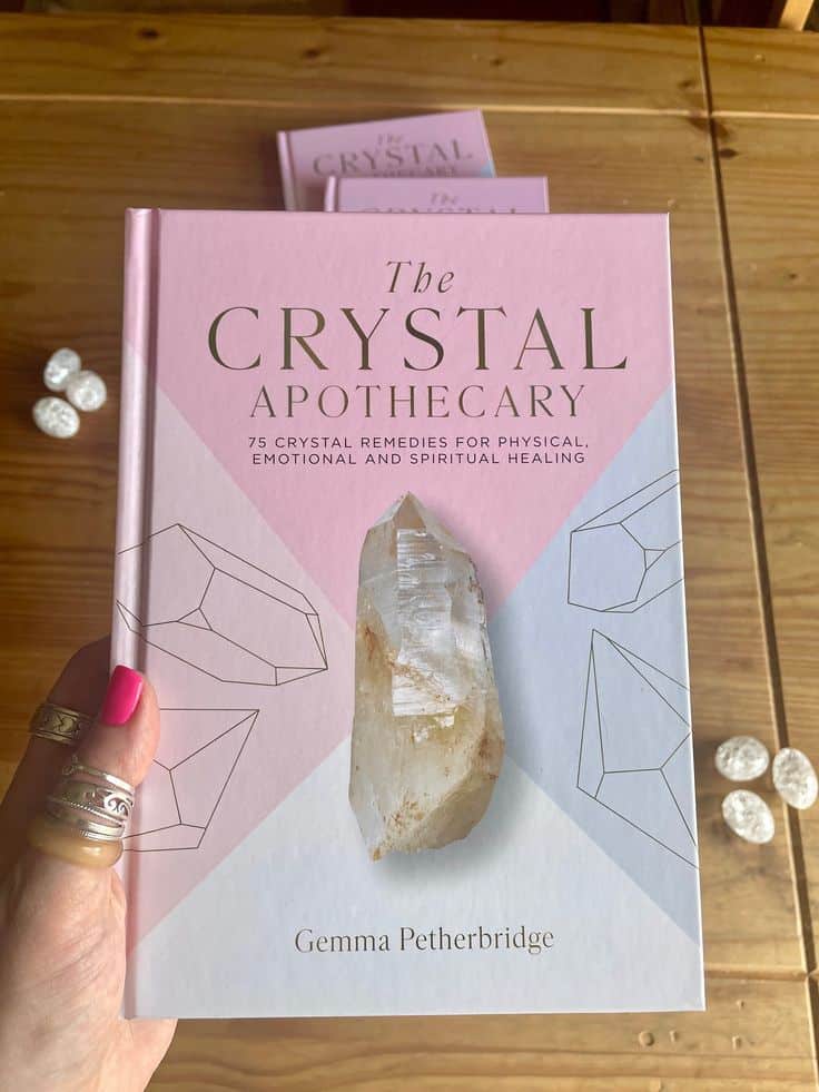 The Crystal Apothecary book held in hand on wooden table with decorative stones nearby.