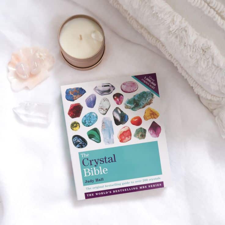 The Crystal Bible book on a bed with crystals, candle, and cozy blankets for a soothing, relaxing atmosphere.