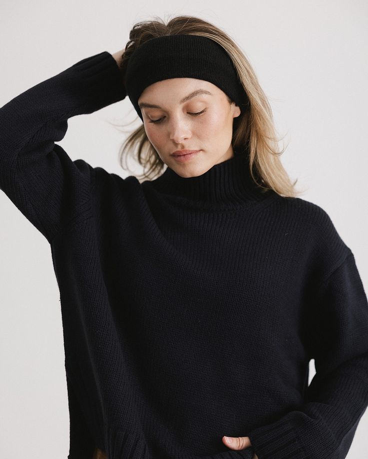 Woman in black sweater and headband posing with eyes closed against a plain background.