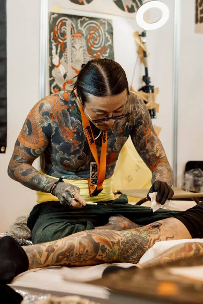 The highlights of the great Gods of Ink Tattoo Convention 2025 - Tattoo Life Tattoo artist creating detailed body art with traditional technique at a tattoo convention. | Sky Rye Design Tattoo artist creating detailed body art with traditional technique at a tattoo convention.