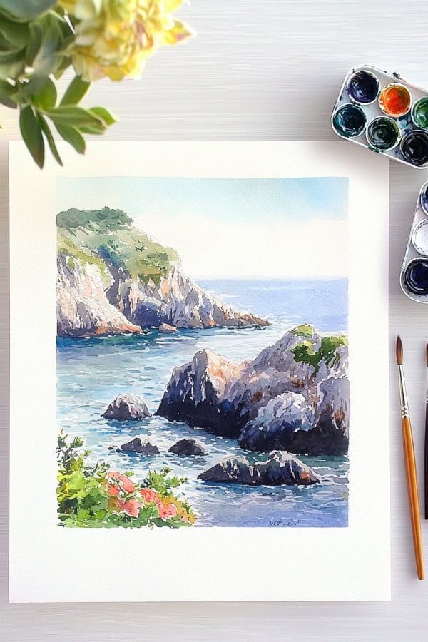 Watercolor painting of a coastal landscape with rocky cliffs, surrounded by paintbrushes and a watercolor palette.