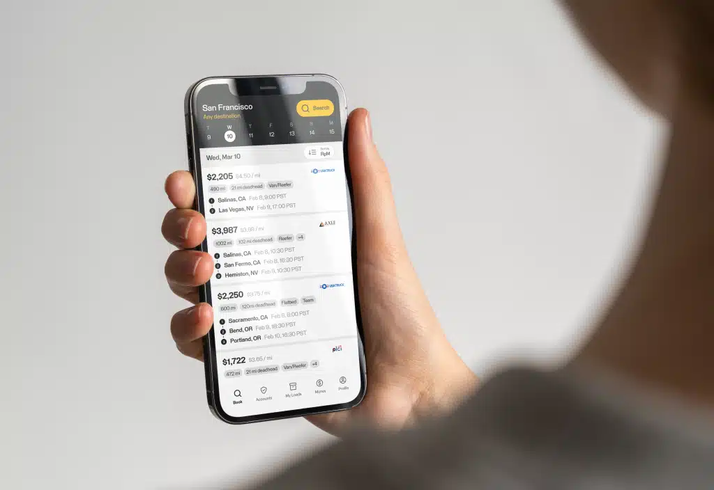 TruckSmarter-Mockup-HomeScreen-1 Person holding a smartphone displaying a transportation app with a list of freight rates and destinations. | Sky Rye Design Person holding a smartphone displaying a transportation app with a list of freight rates and destinations.