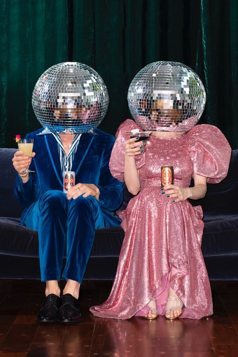Two people in disco ball helmets and colorful outfits sit on a couch, holding drinks and enjoying a party vibe.