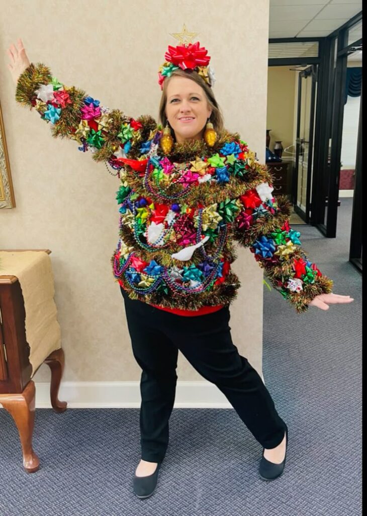 Person in festive, colorful Christmas sweater adorned with tinsel, bows, and decorations, striking a joyful pose.