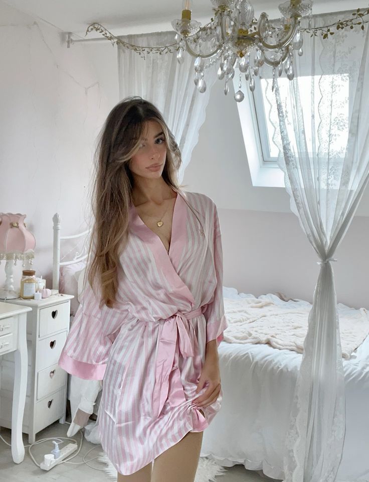 Woman in pink striped robe stands in elegant bedroom with white decor and chandelier.