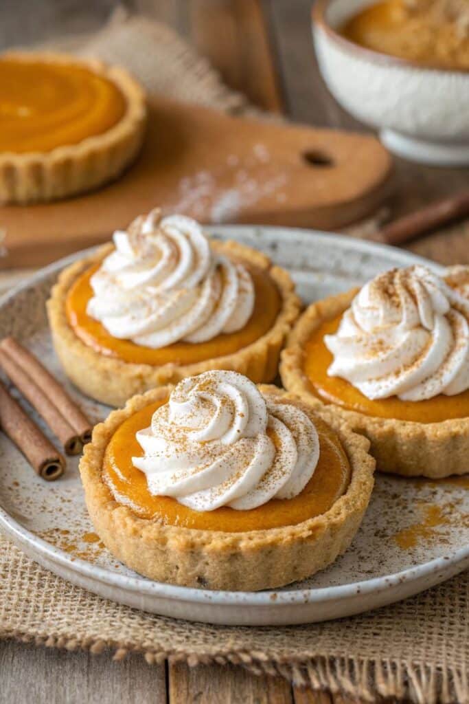 Vegan Pumpkin Pie Cookies Recipe Mini pumpkin pies topped with whipped cream and cinnamon on a rustic plate, surrounded by cinnamon sticks. | Sky Rye Design Mini pumpkin pies topped with whipped cream and cinnamon on a rustic plate, surrounded by cinnamon sticks.