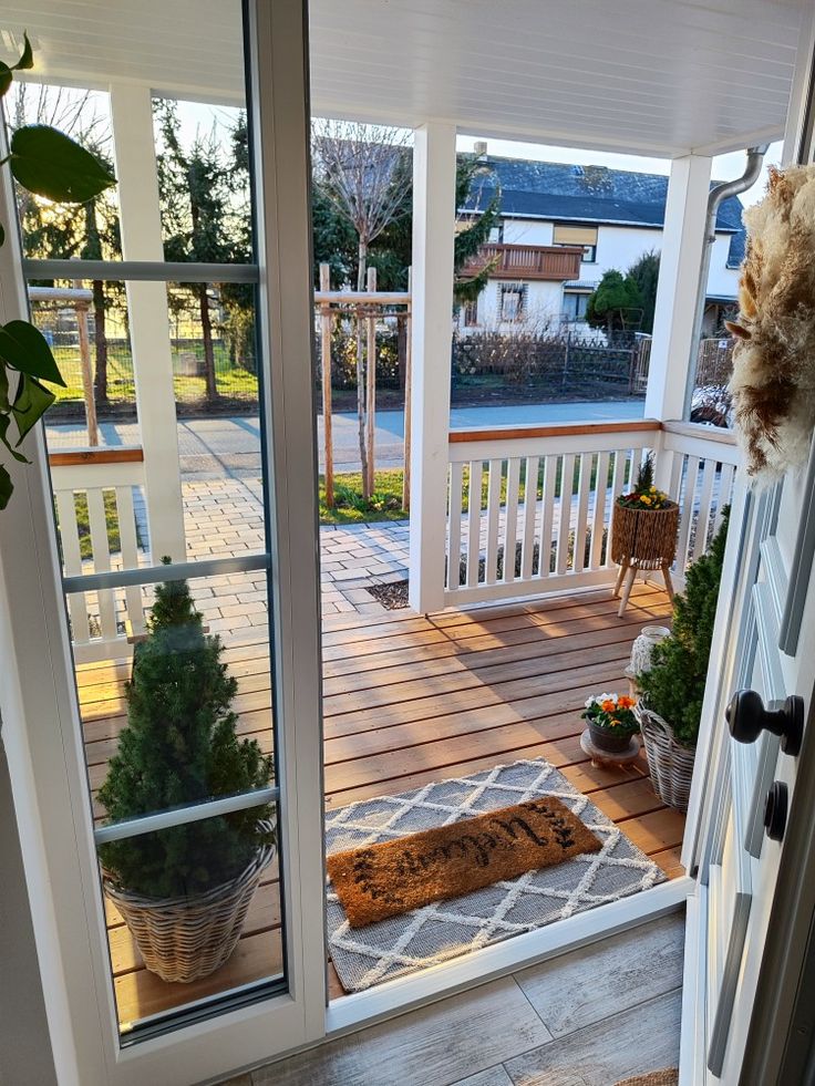 Welcoming front porch view with potted plants, cozy doormat, and sunlight casting shadows, creating a warm entrance.