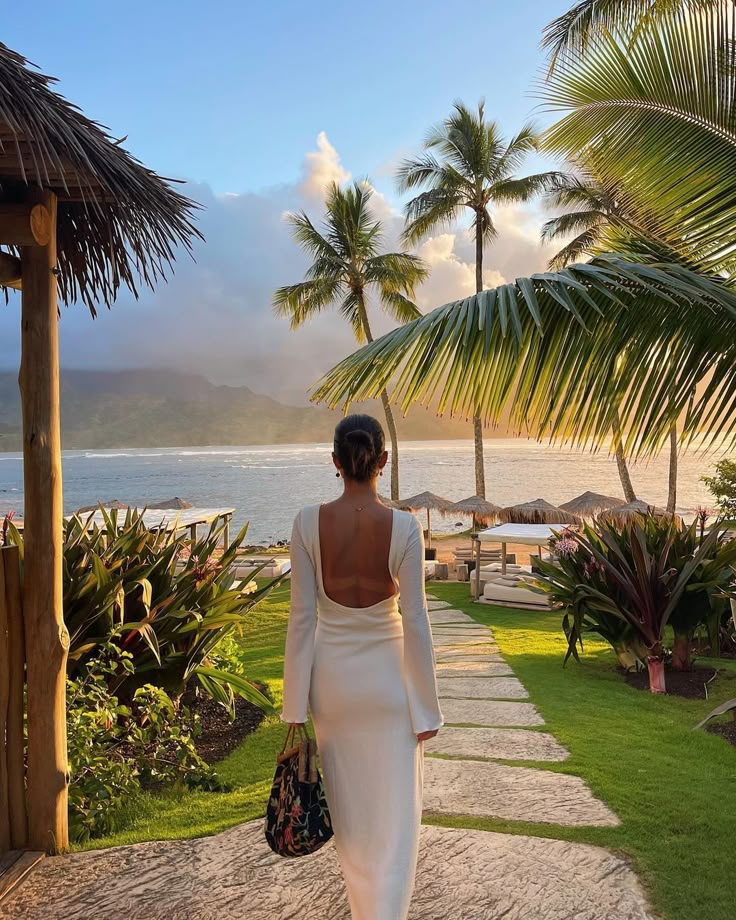 Woman in white dress walking towards tropical beach at sunset, surrounded by palm trees and serene ocean view.