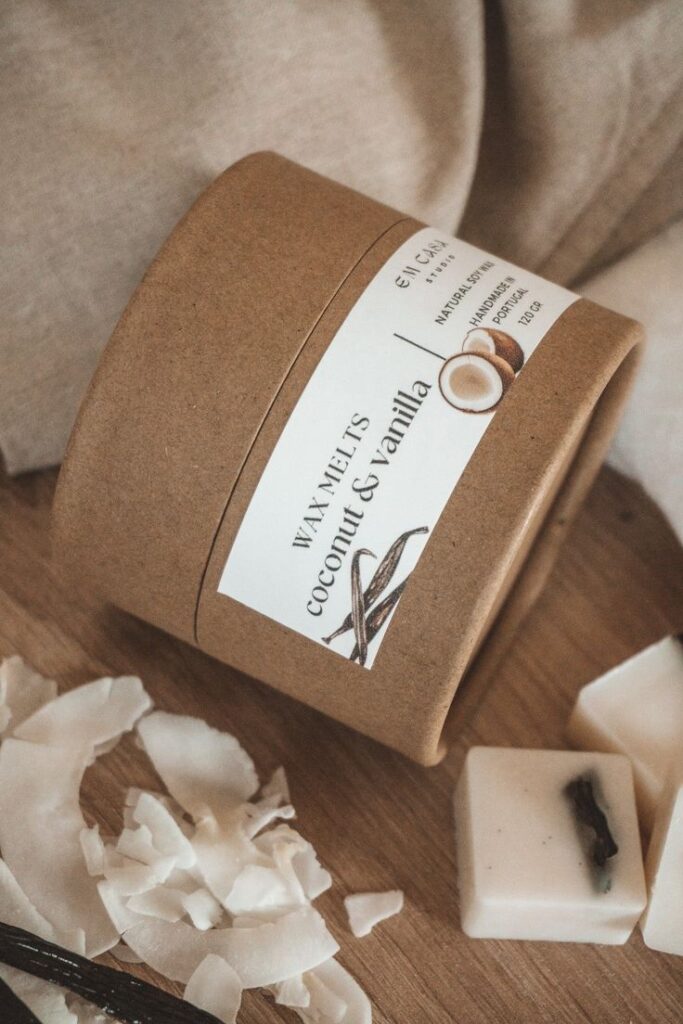 WAX MELT COCO BAUNILHA Brown cardboard container of coconut and vanilla wax melts beside coconut flakes on a wooden surface. | Sky Rye Design Brown cardboard container of coconut and vanilla wax melts beside coconut flakes on a wooden surface.