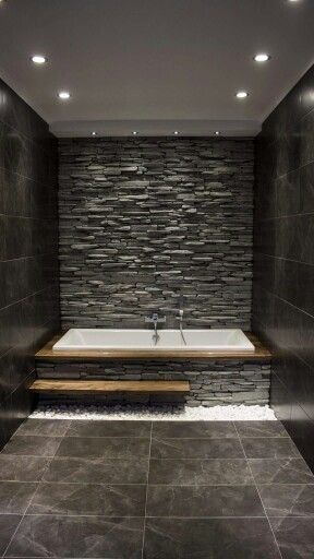 Modern bathroom with stone wall and built-in bathtub, featuring recessed lighting and dark floor tiles for a sleek design.