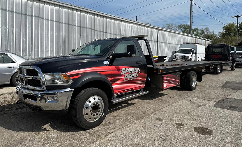 Black flatbed tow truck with Speedy branding parked in an industrial lot on a sunny day.