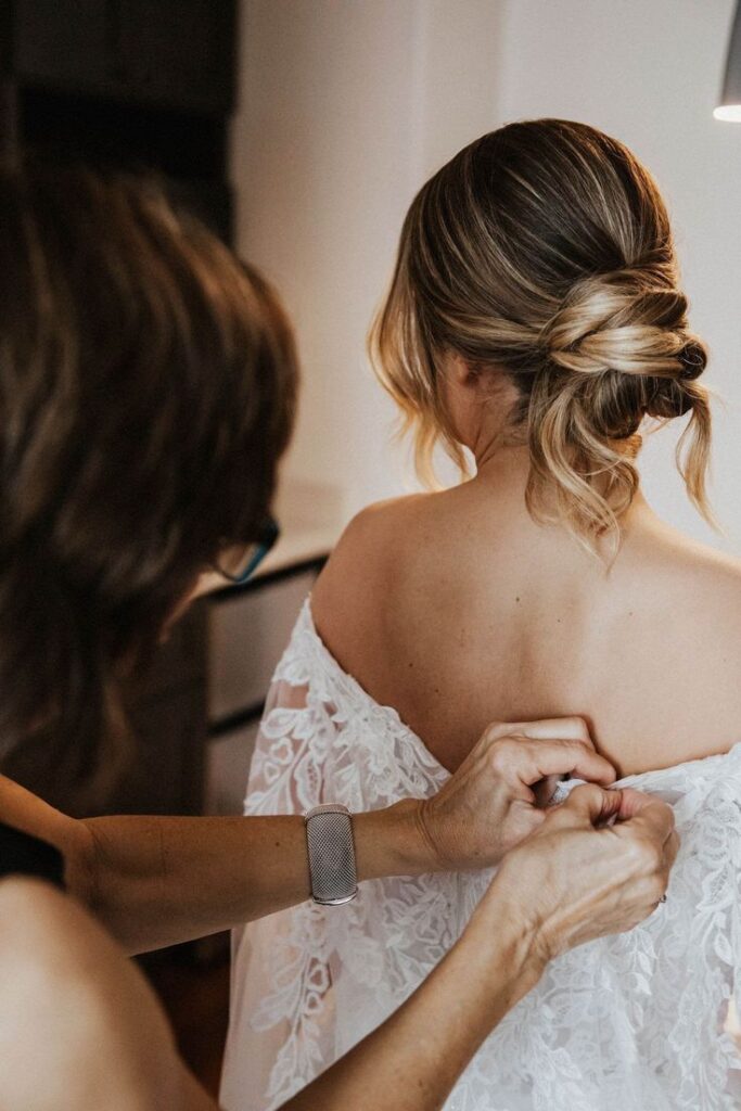 Woman helping bride with lace wedding dress, hair styled in elegant updo.