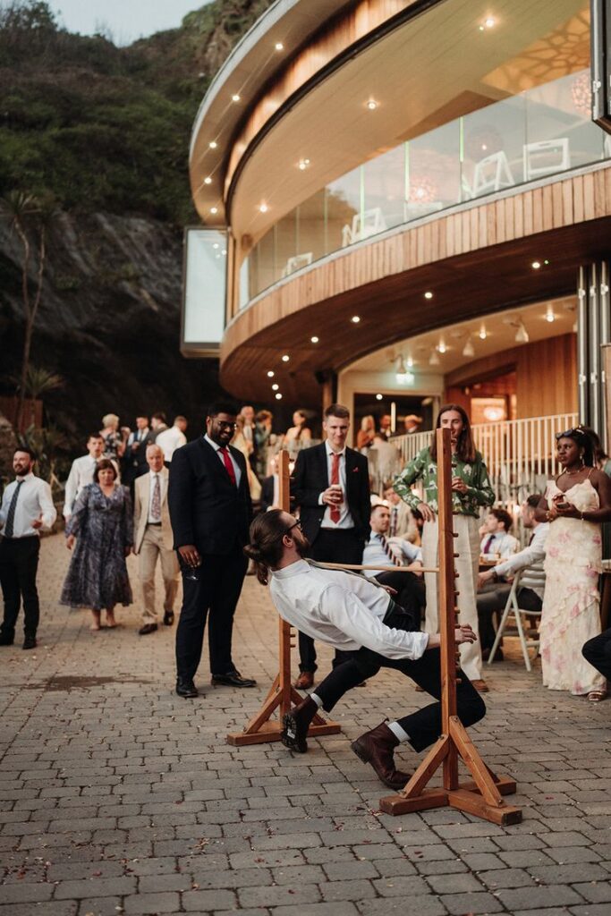 Man in white shirt playing limbo at outdoor party with crowd watching near modern building.