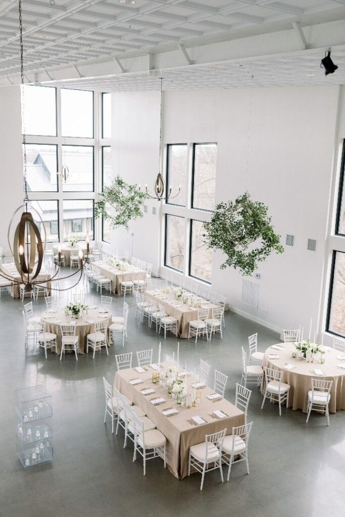 Elegant banquet hall with large windows, round and rectangular tables, white chairs, and green hanging decor. Perfect for events.