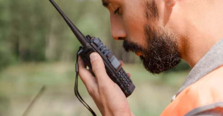 Man in orange vest communicates using a walkie-talkie outdoors in a green, blurred background setting.