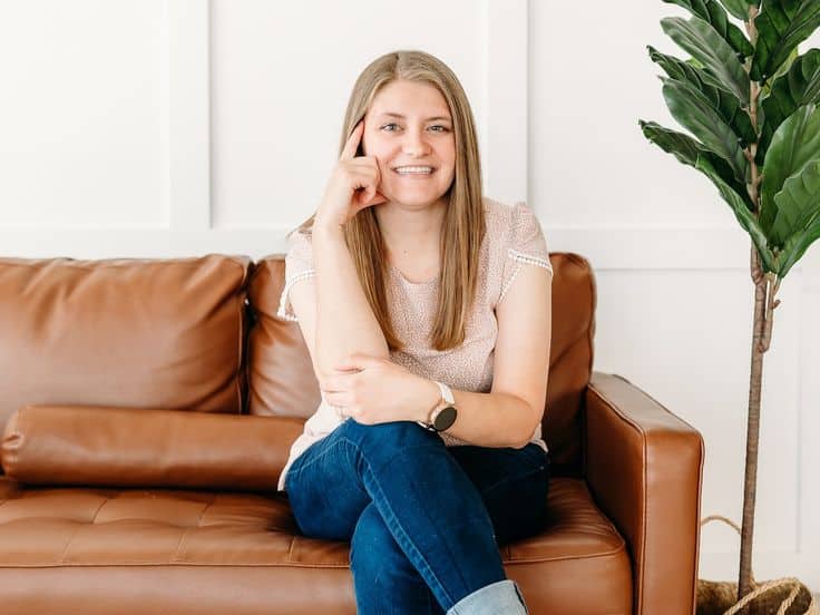 Smiling woman sitting on a brown leather couch with a plant, wearing casual attire, in a bright room.