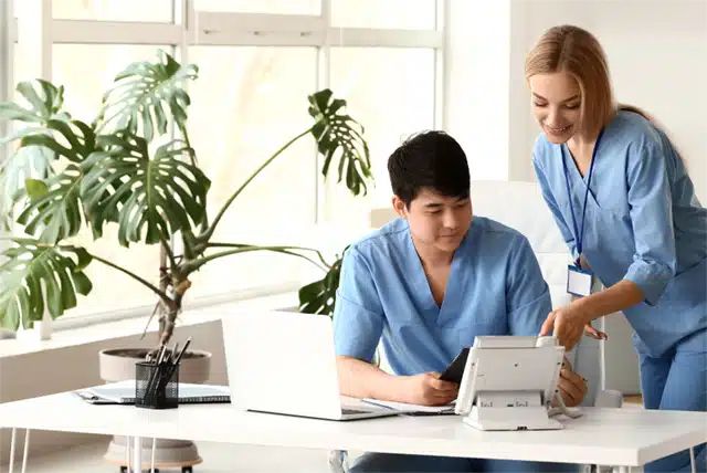 What-does-a-healthcare-assistant-do Two healthcare professionals in scrubs collaborating at a desk with a laptop and tablet in a medical office. | Sky Rye Design Two healthcare professionals in scrubs collaborating at a desk with a laptop and tablet in a medical office.