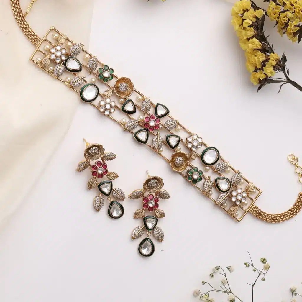 Elegant gold necklace and earrings with colorful gemstones and floral design, displayed with yellow flowers.