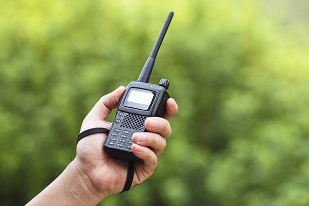 Hand holding a two-way radio with an antenna, outdoors with greenery in the background.
