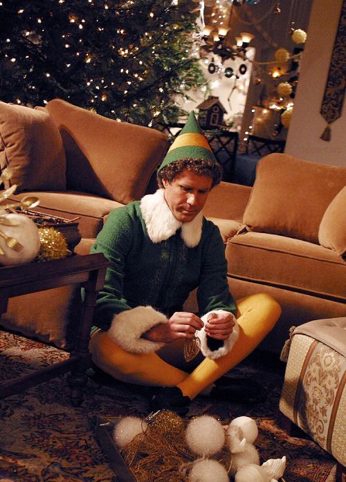 Will Ferrell as Buddy Hobbs aka _Buddy the Elf_ in Elf 2003 Man in green elf costume sits on the floor, decorating for Christmas with a festive tree in the background. | Sky Rye Design Man in green elf costume sits on the floor, decorating for Christmas with a festive tree in the background.