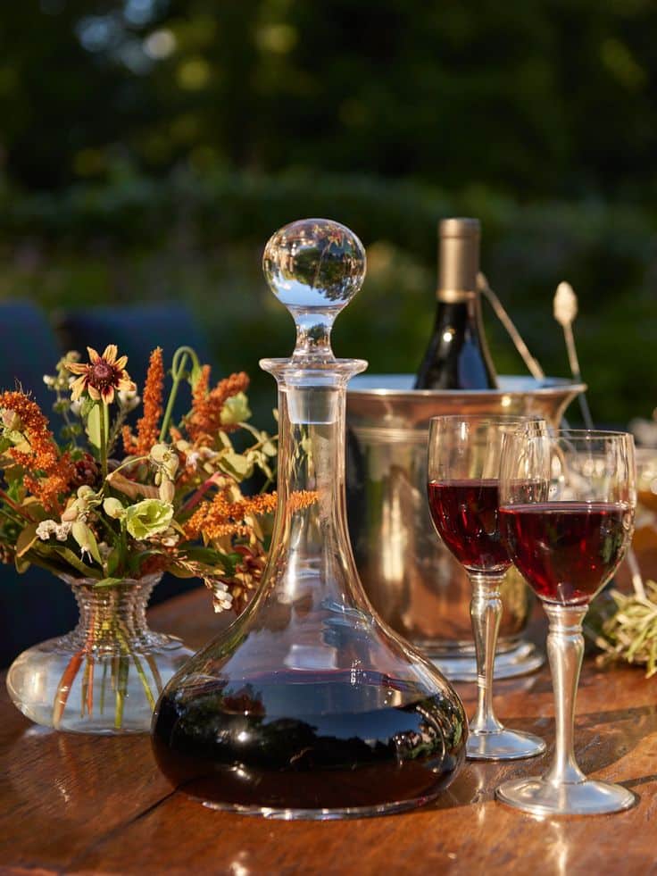 William Yeoward Crystal Classic Ships Decanter Decanter and wine glasses on rustic table, set for an elegant outdoor gathering with flowers and wine bottle background. | Sky Rye Design Decanter and wine glasses on rustic table, set for an elegant outdoor gathering with flowers and wine bottle background.