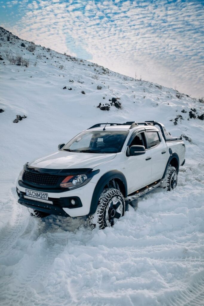 Winter Driving Four-wheel drive white pickup truck navigating snowy mountain terrain under a clear blue sky. | Sky Rye Design Four-wheel drive white pickup truck navigating snowy mountain terrain under a clear blue sky.