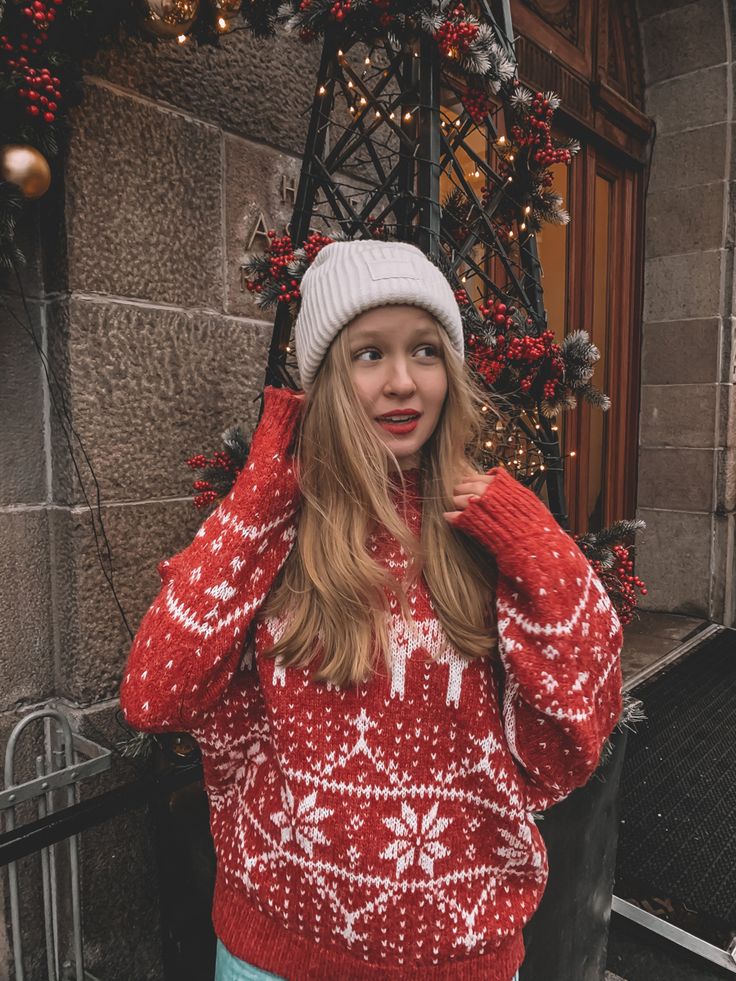 Winter mood Woman in festive red sweater and white beanie, standing by holiday decorations outside a building. | Sky Rye Design Woman in festive red sweater and white beanie, standing by holiday decorations outside a building.