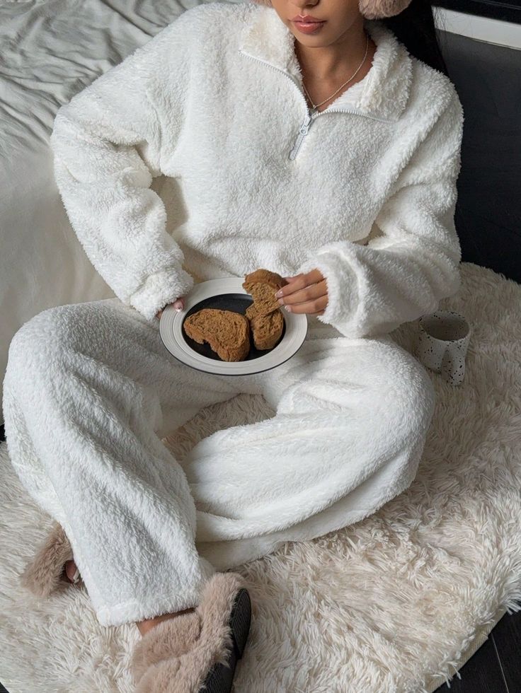 Cozy in white fleece loungewear with earmuffs, enjoying cookies on a fluffy rug.