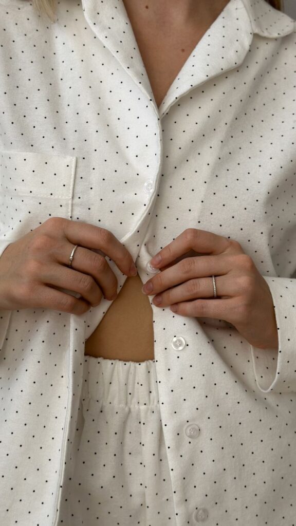 Person buttoning a white polka dot pajama shirt, showcasing minimalist silver rings.