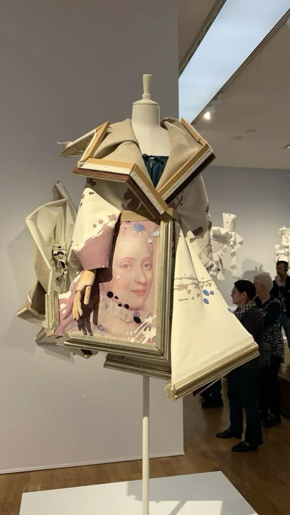 Dress sculpture with art frames and fabric, featuring a painted face, displayed in a modern gallery setting.
