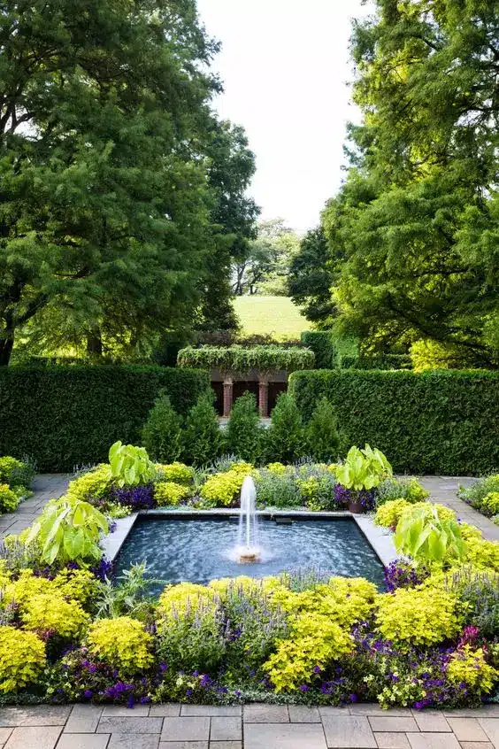 a-large-cool-modern-fountain-surrounded-with-bold-blooms-and-greenery-is-a-fantastic-idea-for-a-modern-outdoor-space Elegant garden with a central fountain, vibrant flowers, and lush greenery surrounded by tall trees in a serene landscape. | Sky Rye Design Elegant garden with a central fountain, vibrant flowers, and lush greenery surrounded by tall trees in a serene landscape.