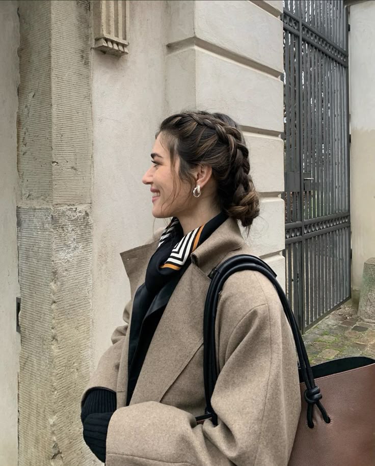 Stylish woman in a beige coat with braided hair, smiling near a stone wall and iron gate.