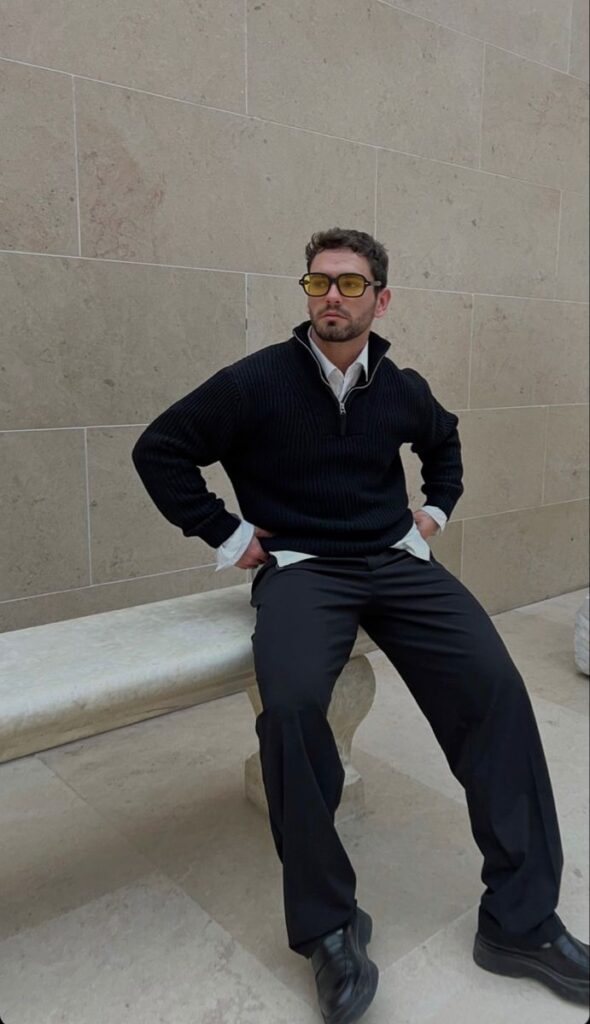 Man in black sweater and sunglasses seated on a stone bench in a minimalist setting.
