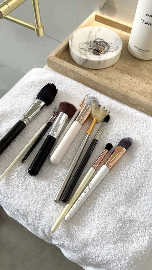 a6983b6e-7ff4-42a2-977a-f3202c45d8db A collection of makeup brushes on a white towel, next to a marble accessory tray and hand lotion. | Sky Rye Design A collection of makeup brushes on a white towel, next to a marble accessory tray and hand lotion.