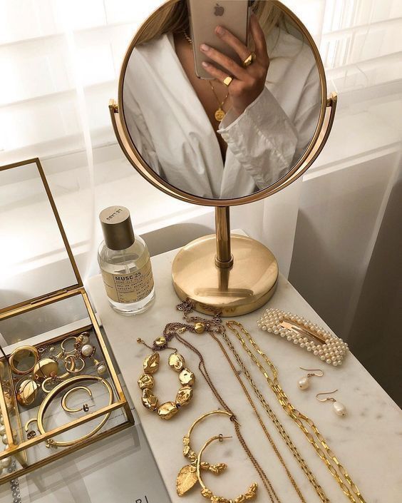 Vanity table with gold jewelry, perfume, and a round mirror reflecting a person wearing a white shirt, showcasing elegance.