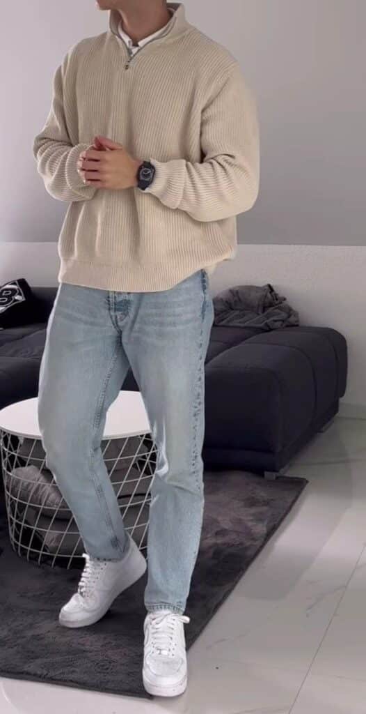 Casual outfit featuring beige sweater, light jeans, and white sneakers in a modern living room setting.