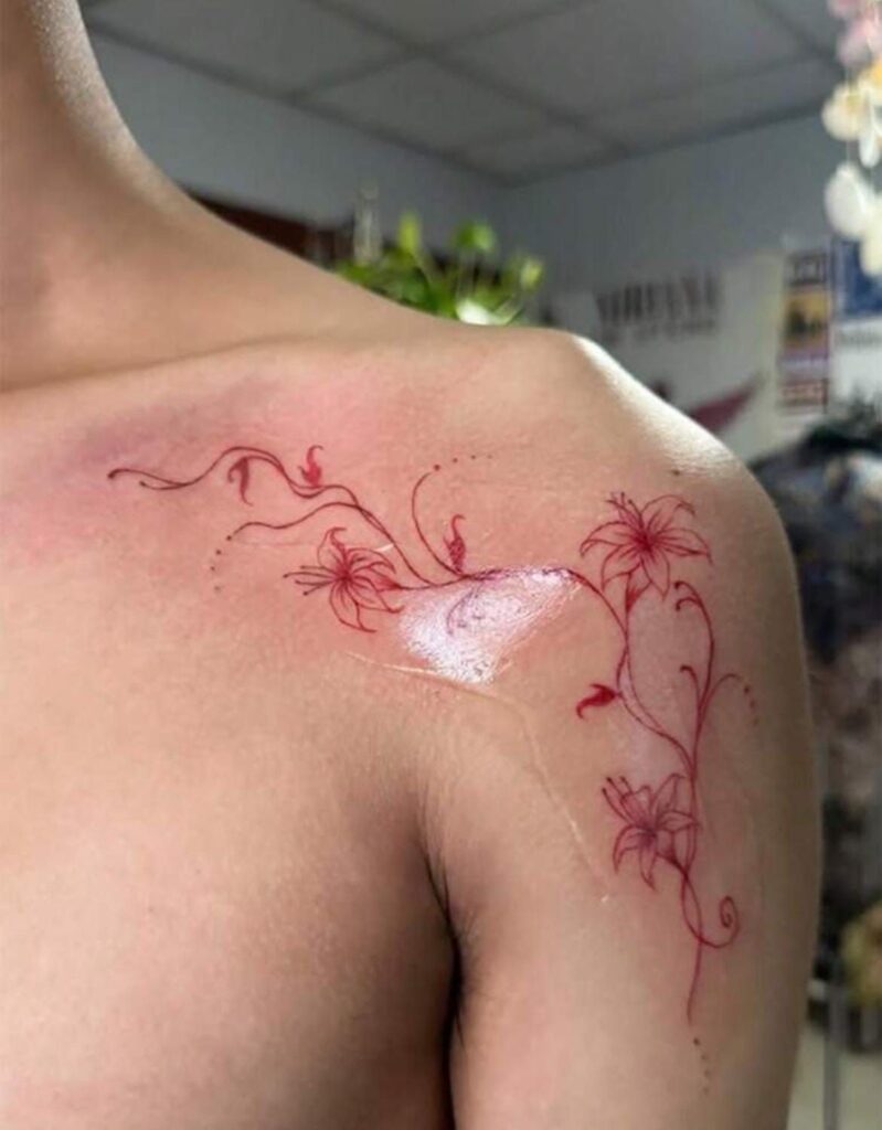 Delicate red floral tattoo on shoulder, showcasing intricate vine design with blooming flowers, set in a well-lit room.