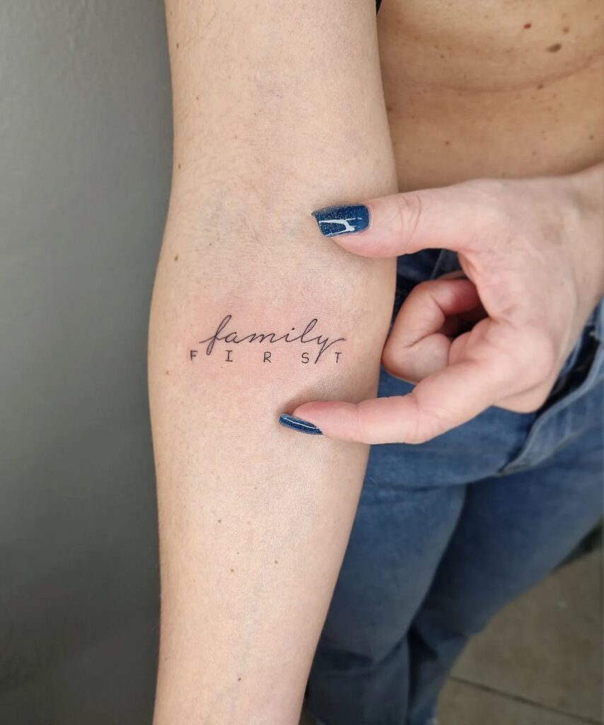 Arm tattoo reading family first with blue-polished nails pointing at it.