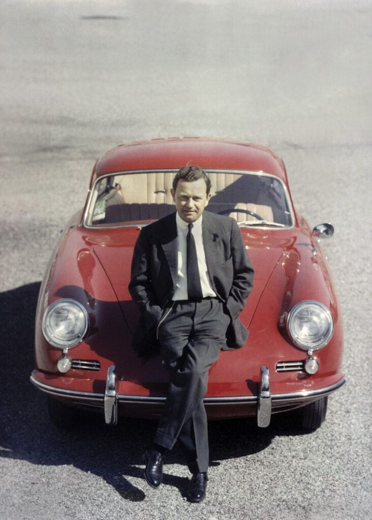 Man in suit leaning on a classic red Porsche, parked on a sunny day, vintage style.