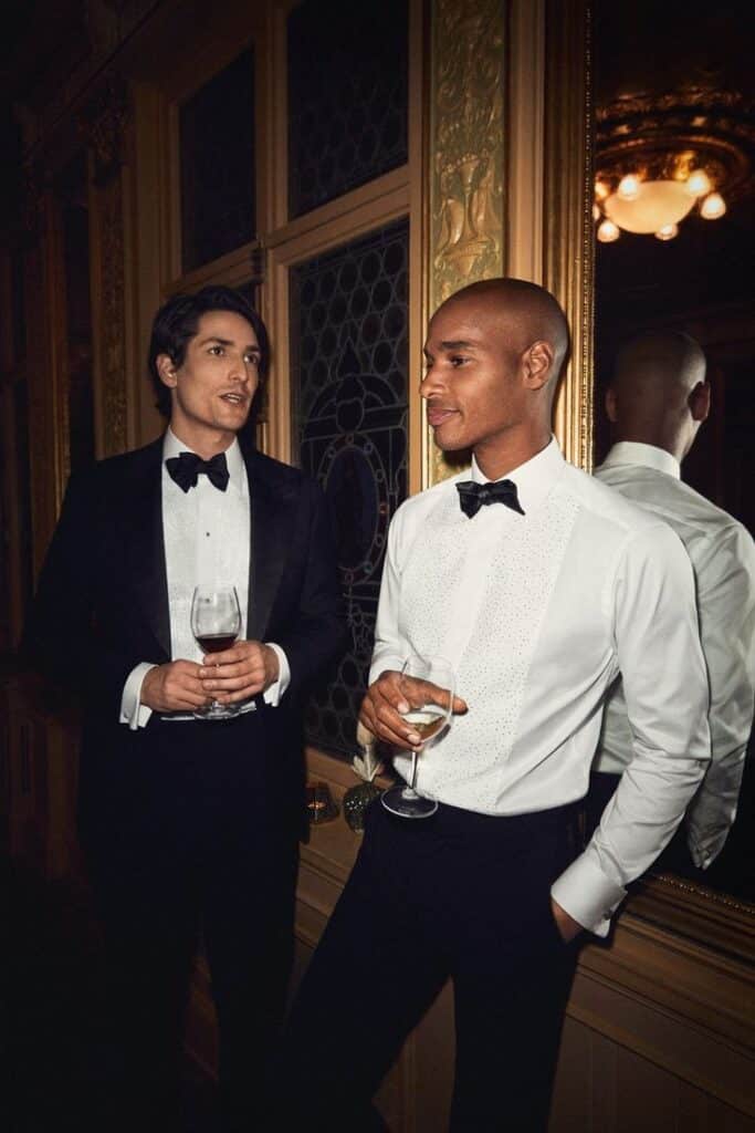 Two men in elegant tuxedos socializing with wine at a formal evening event, reflecting in a mirror.