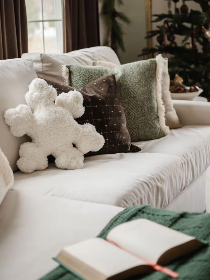Screenshot Cozy living room with festive pillows and a plush snowflake on the couch near a Christmas tree and open book. | Sky Rye Design Cozy living room with festive pillows and a plush snowflake on the couch near a Christmas tree and open book.