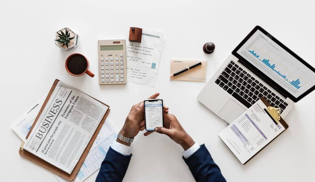 Business desk setup with newspaper, laptop, calculator, documents, and phone in hand for financial analysis.