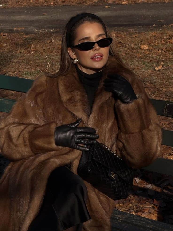 Stylish woman in fur coat, sunglasses, and gloves sits on a bench, holding a quilted handbag in a fall setting.