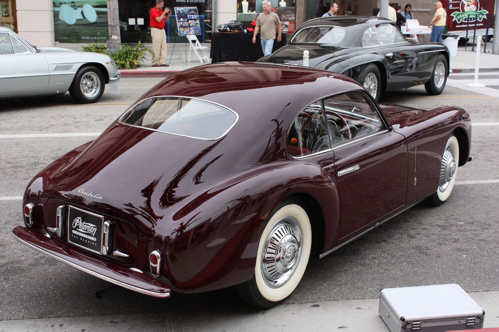 Classic burgundy sports car displayed at a vintage car show on a city street.
