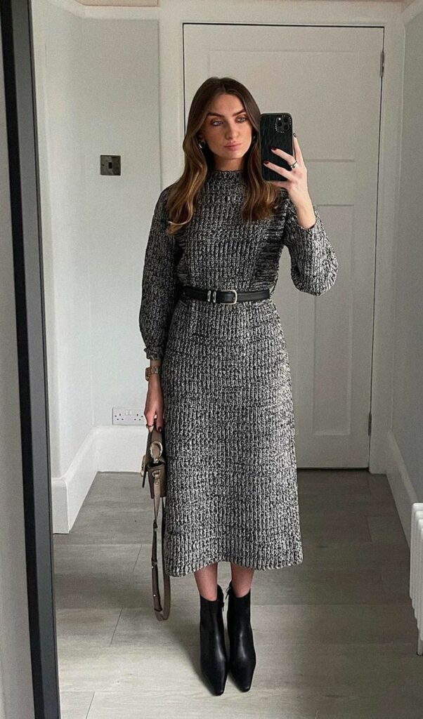 Woman in a gray knit dress with belt, taking a mirror selfie, wearing black ankle boots. Stylish outfit in minimal room.