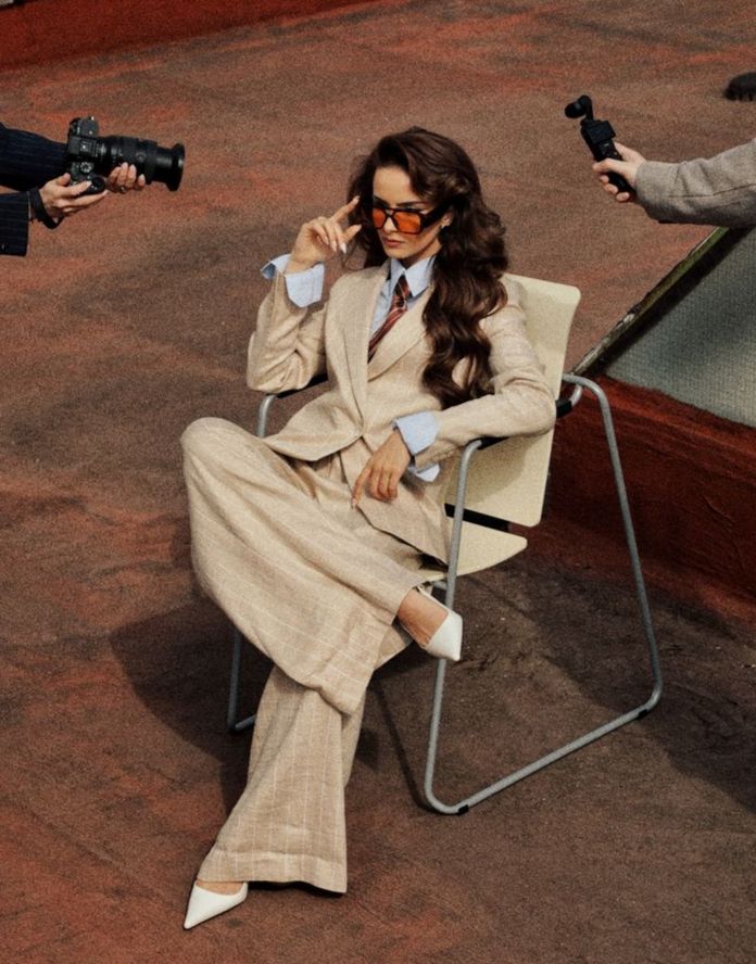 Woman in beige suit and sunglasses sitting on a chair, surrounded by photographers capturing the scene.