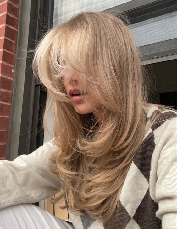 Stylish woman with long, layered blonde hair and a cozy diamond-patterned sweater gazes outdoors.